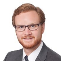 A man with light skin, short red hair, a beard, and glasses is wearing a gray suit, white shirt, and striped tie, posed against a plain white background.