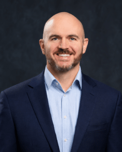A man with a shaved head and trimmed beard wearing a blue dress shirt and navy suit jacket smiles against a dark, plain background.