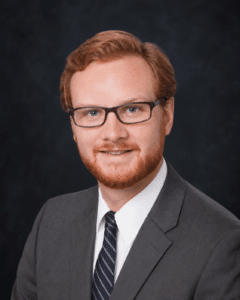 A man with red hair, a beard, and glasses wearing a gray suit, white shirt, and striped tie poses for a formal studio portrait against a dark background.