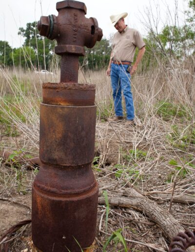 A man in a cowboy hat stands in dry grass, looking at a rusted, capped wellhead in the foreground.