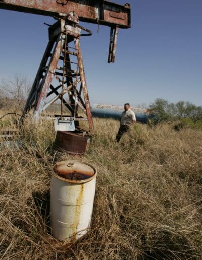 A man stands in tall grass near a rusty oil pumpjack and barrels under a clear sky.